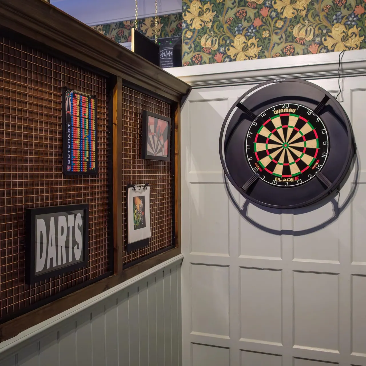 Darts area with electronic scoreboard and dartboard mounted on white panelled wall, decorated with floral wallpaper above.