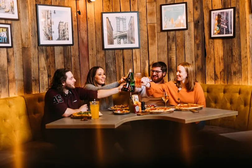 Four friends raise bottles in a toast at a booth with pizza and drinks, wooden walls and framed prints behind them.