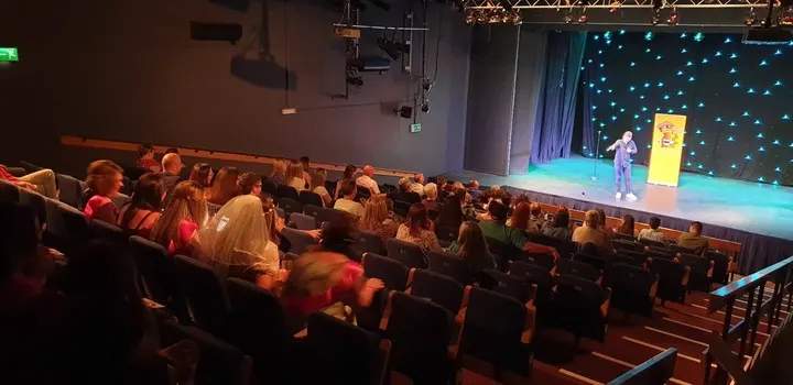 Audience members seated in a theatre watching a performer on an illuminated stage with a starlit backdrop.