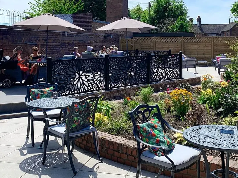 Outdoor patio seating area with guests dining under umbrellas, decorative screens and flowering borders on a sunny day.