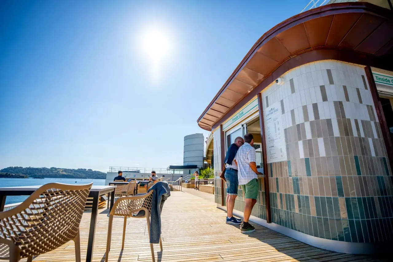 Visitors queue at a waterfront café kiosk on a sunny day, with outdoor seating and blue water visible beyond.