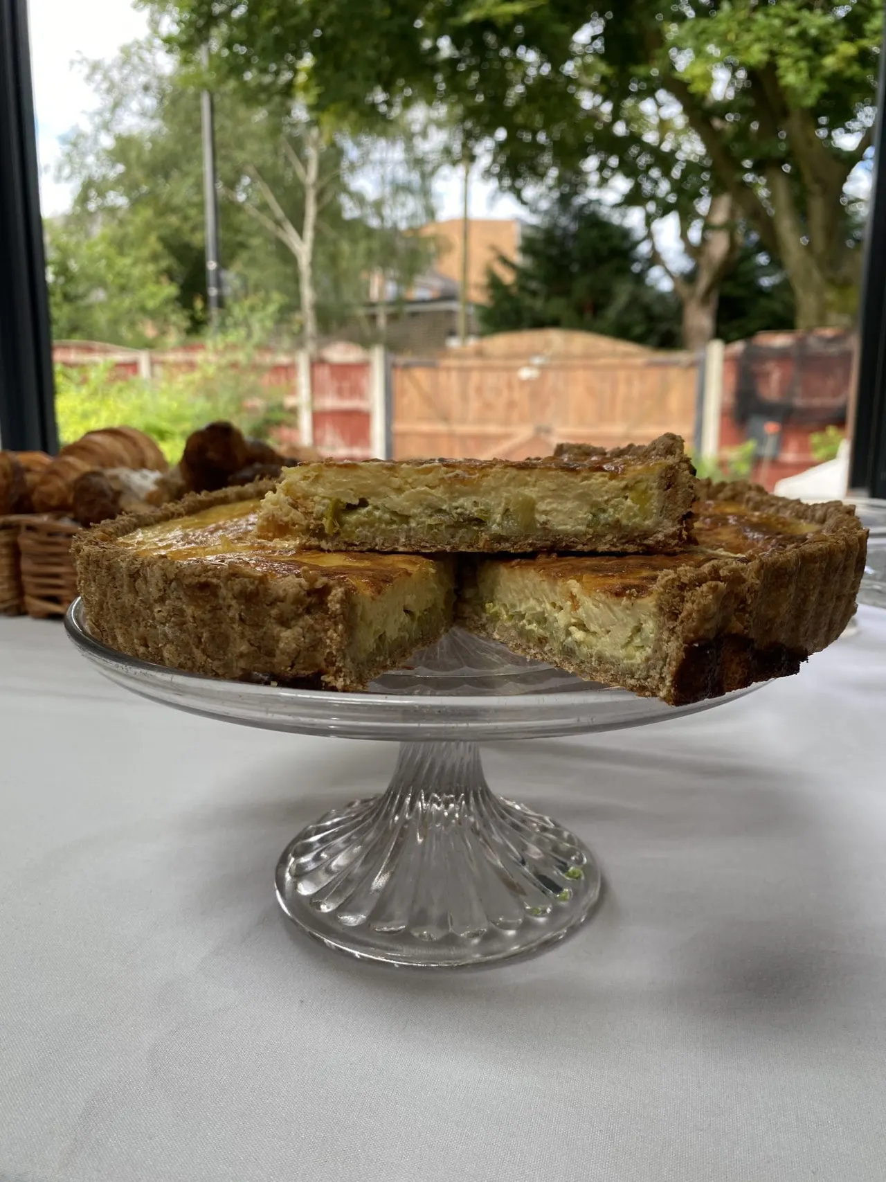 Sliced cake on a glass cake stand displayed by a window overlooking a garden with trees and farm buildings.