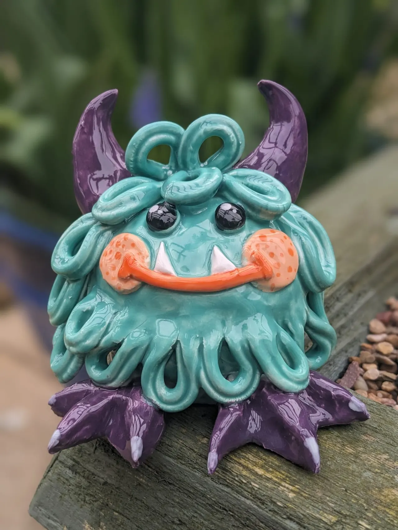 Colourful ceramic creature figurine with turquoise body, purple horns and fins, and orange mouth on a wooden surface outdoors.