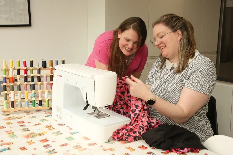 Two young women smiling whilst using a sewing machine at a craft table with fabric and thread spools.