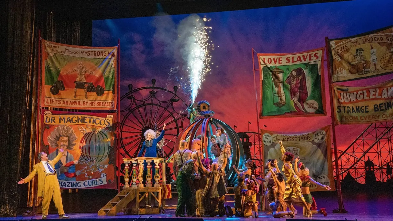 Theatrical stage production featuring performers in period costume around a carnival-themed set with vintage sideshow banners and a ferris wheel prop, with fireworks bursting above.