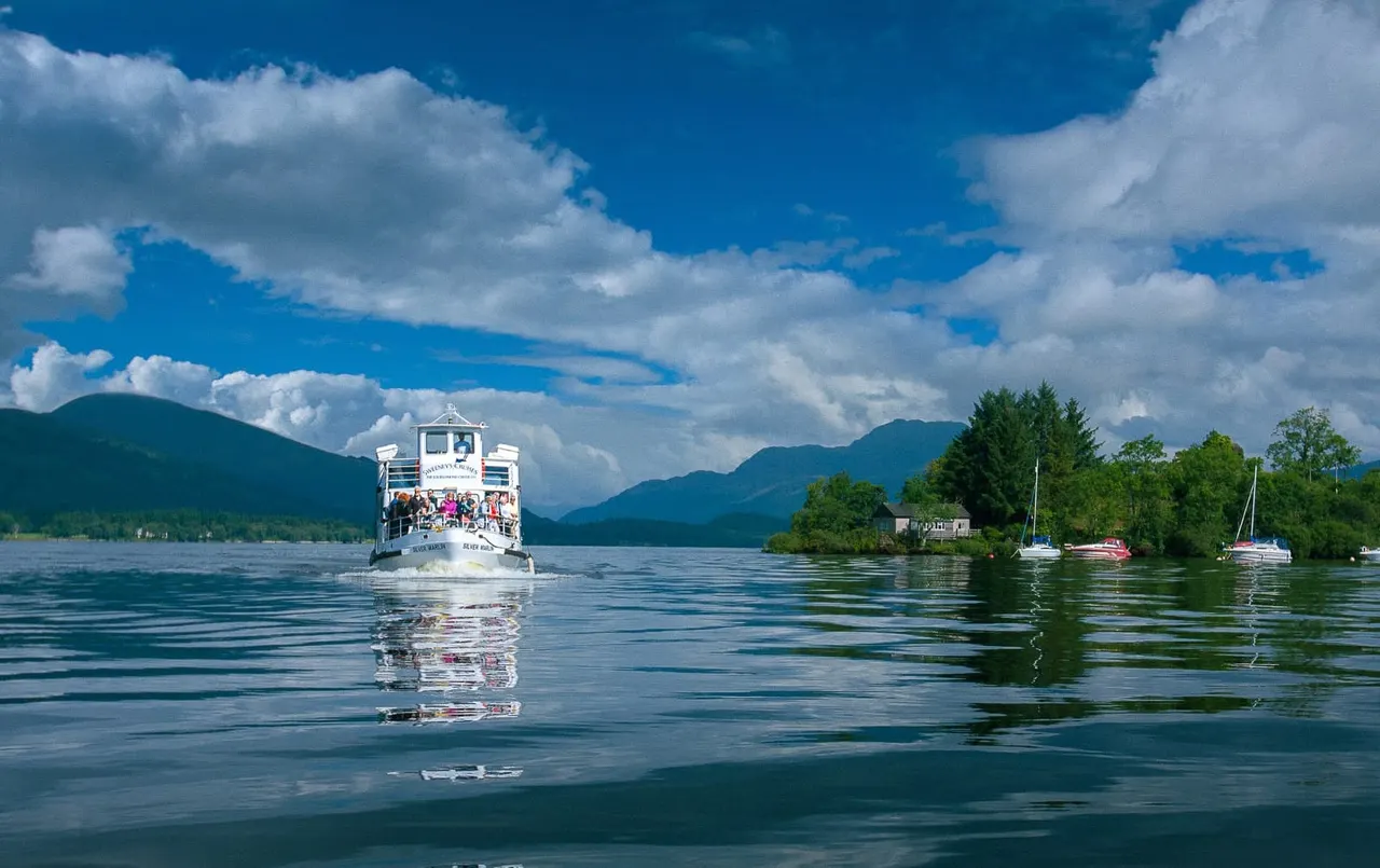 White passenger cruise boat on calm lake water with mountains and forested shores under blue sky with white clouds.