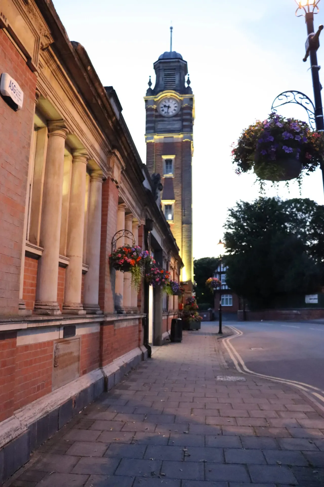 Historic brick town hall with columns beside a street lined with hanging flower baskets, clock tower illuminated in background at dusk.