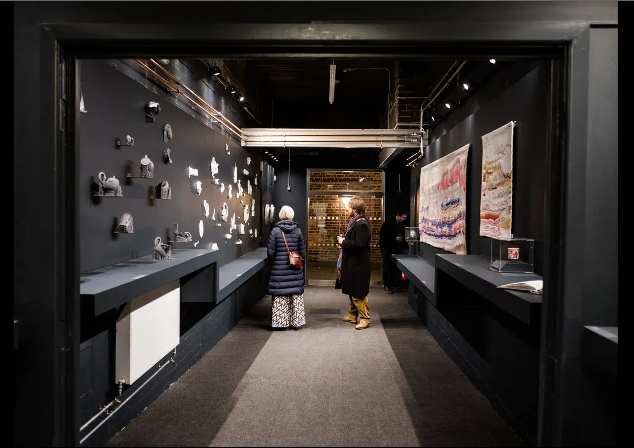 Two visitors explore a museum gallery displaying pottery and ceramics on dark walls and display cases.