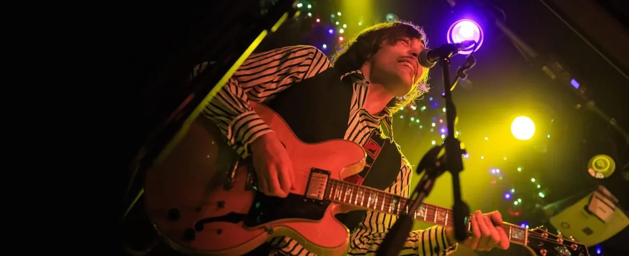 Musician performing with electric guitar on stage under colourful stage lighting and microphone.
