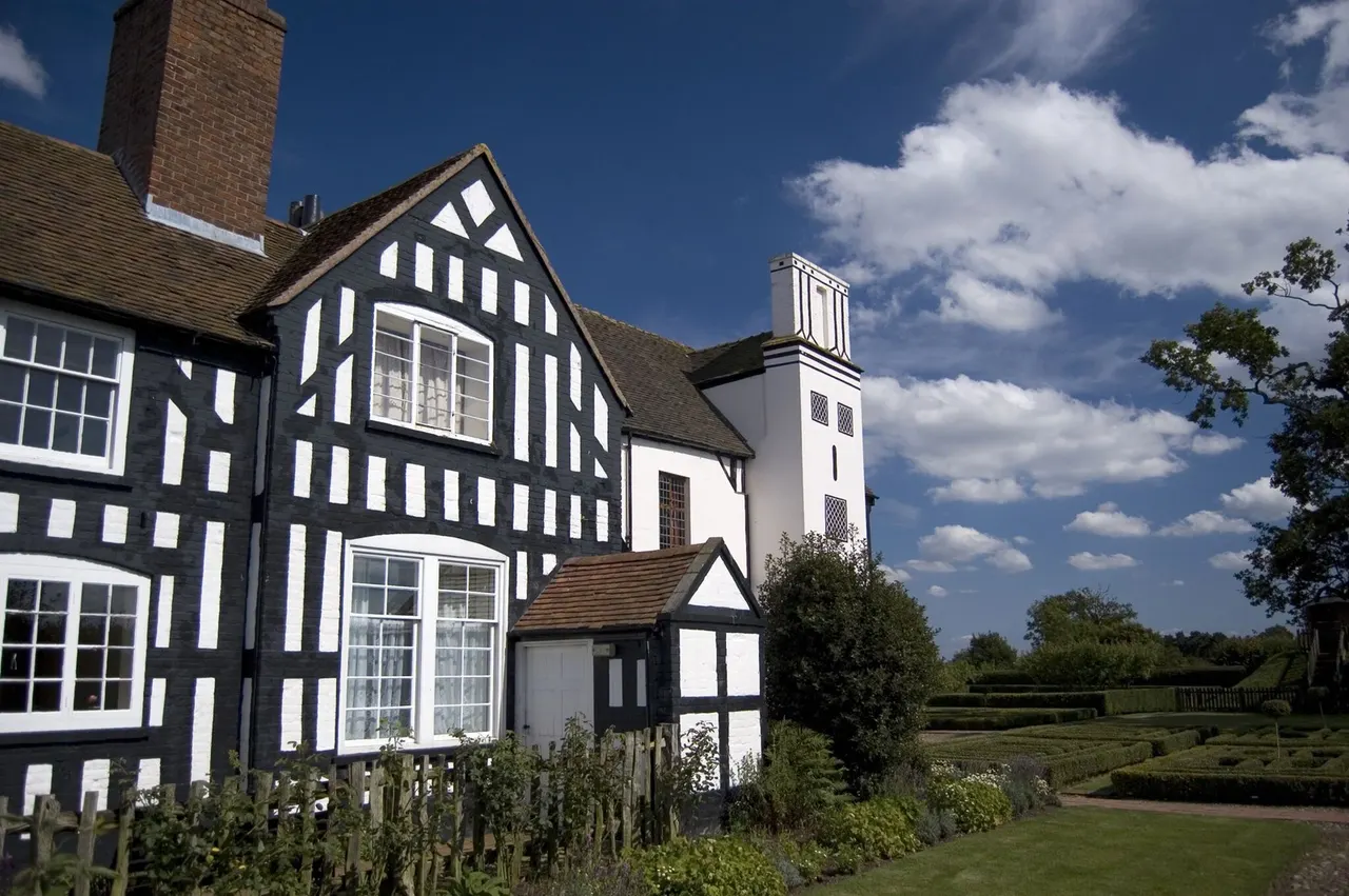 Tudor black-and-white half-timbered house with brick chimney and adjoining church under blue sky with white clouds.