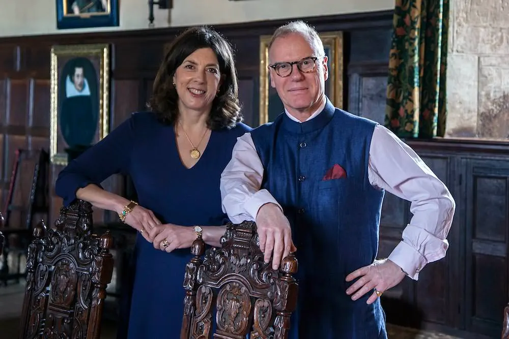 Two people pose together in a historic interior beside ornate carved wooden panels.
