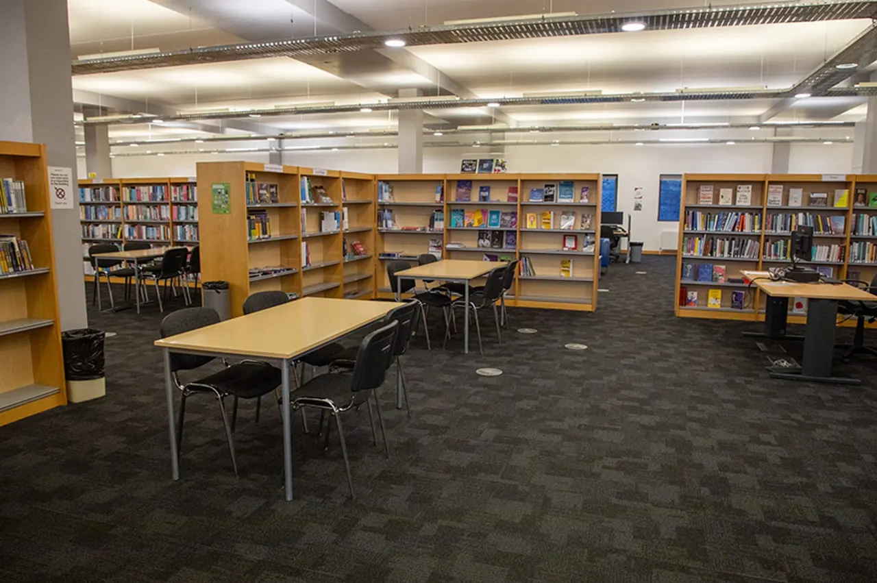 Library interior with wooden bookshelves, study tables and chairs arranged on a dark carpet beneath bright ceiling lights.