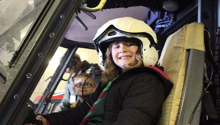 A child wearing a white helmet smiles inside a helicopter cockpit alongside another child.