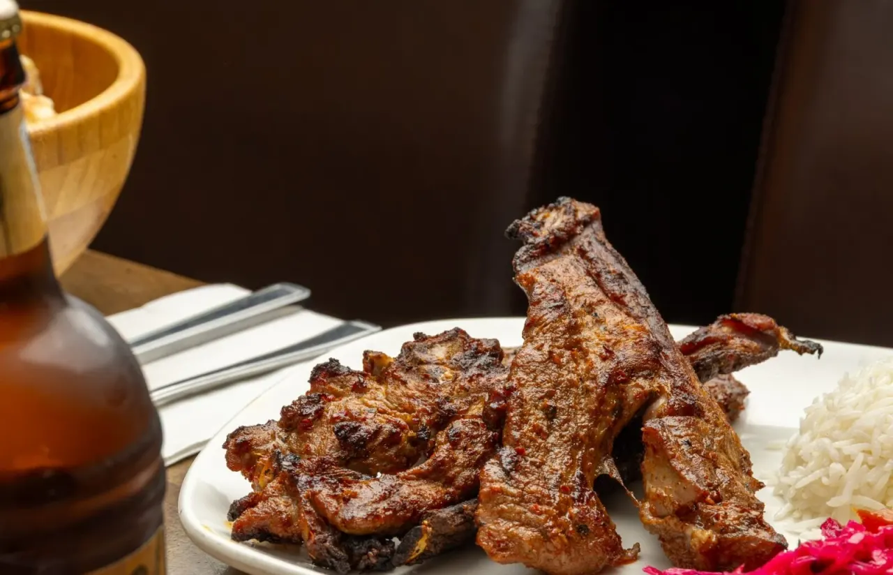 Grilled meat skewers with rice and garnish on a white plate, with a drink and wooden utensils.