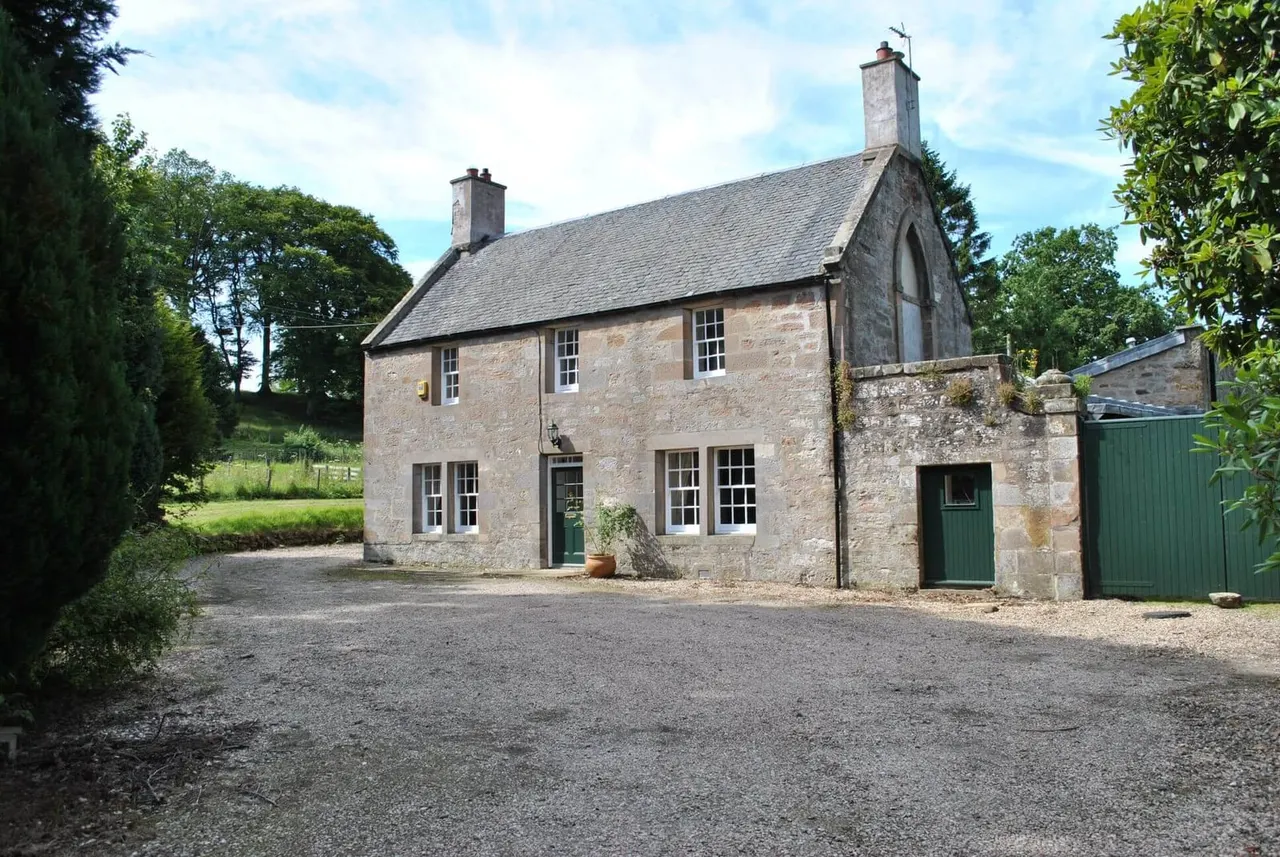 A two-storey stone cottage with slate roof surrounded by trees and green grounds, featuring twin chimneys and green doors.