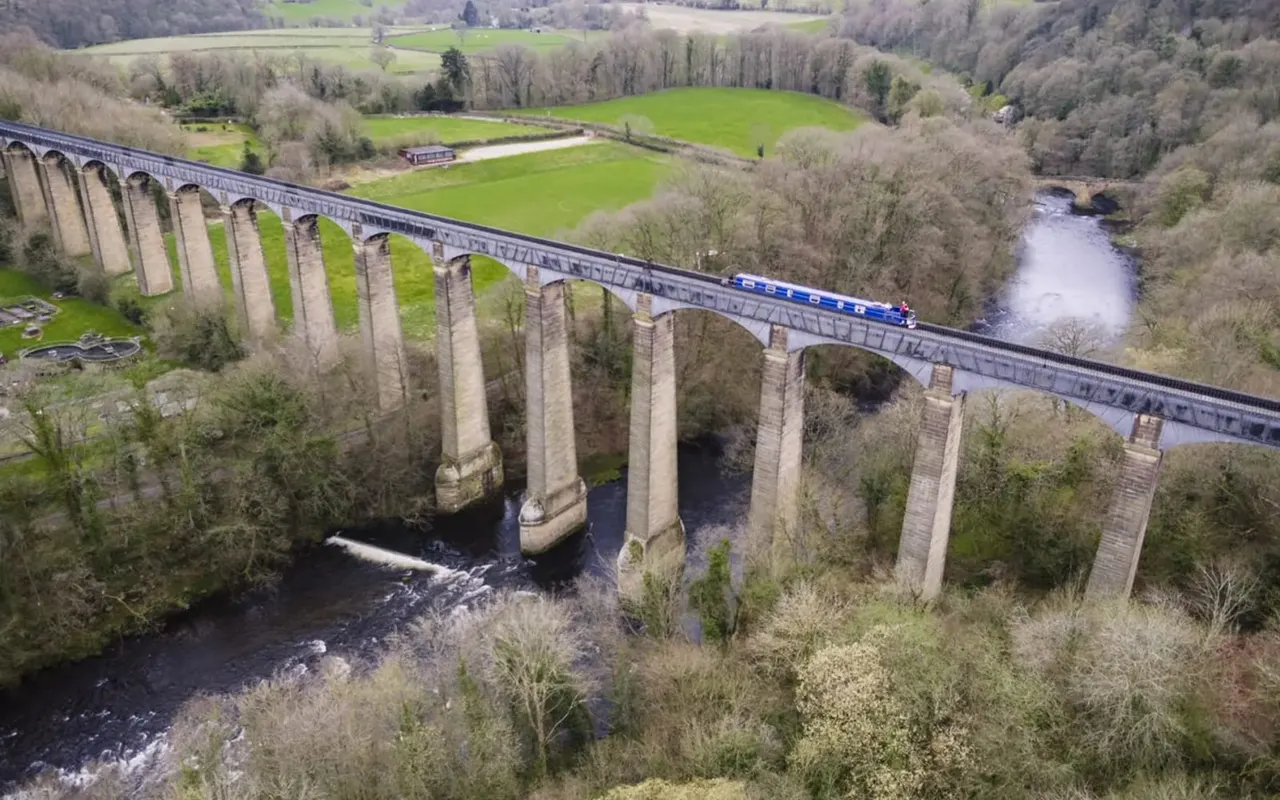A blue passenger train crosses a multi-arched stone viaduct over a river in rural countryside with fields and bare trees.