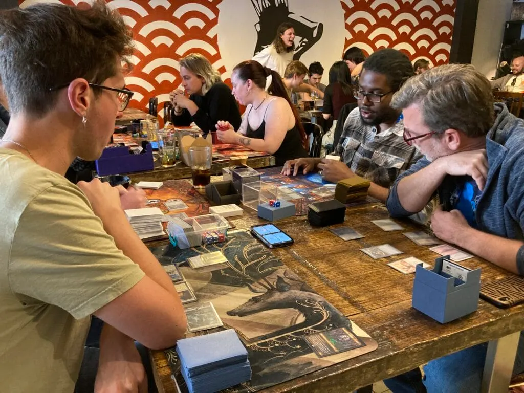 Groups of people playing trading card games at tables in a gaming venue with geometric wall art.