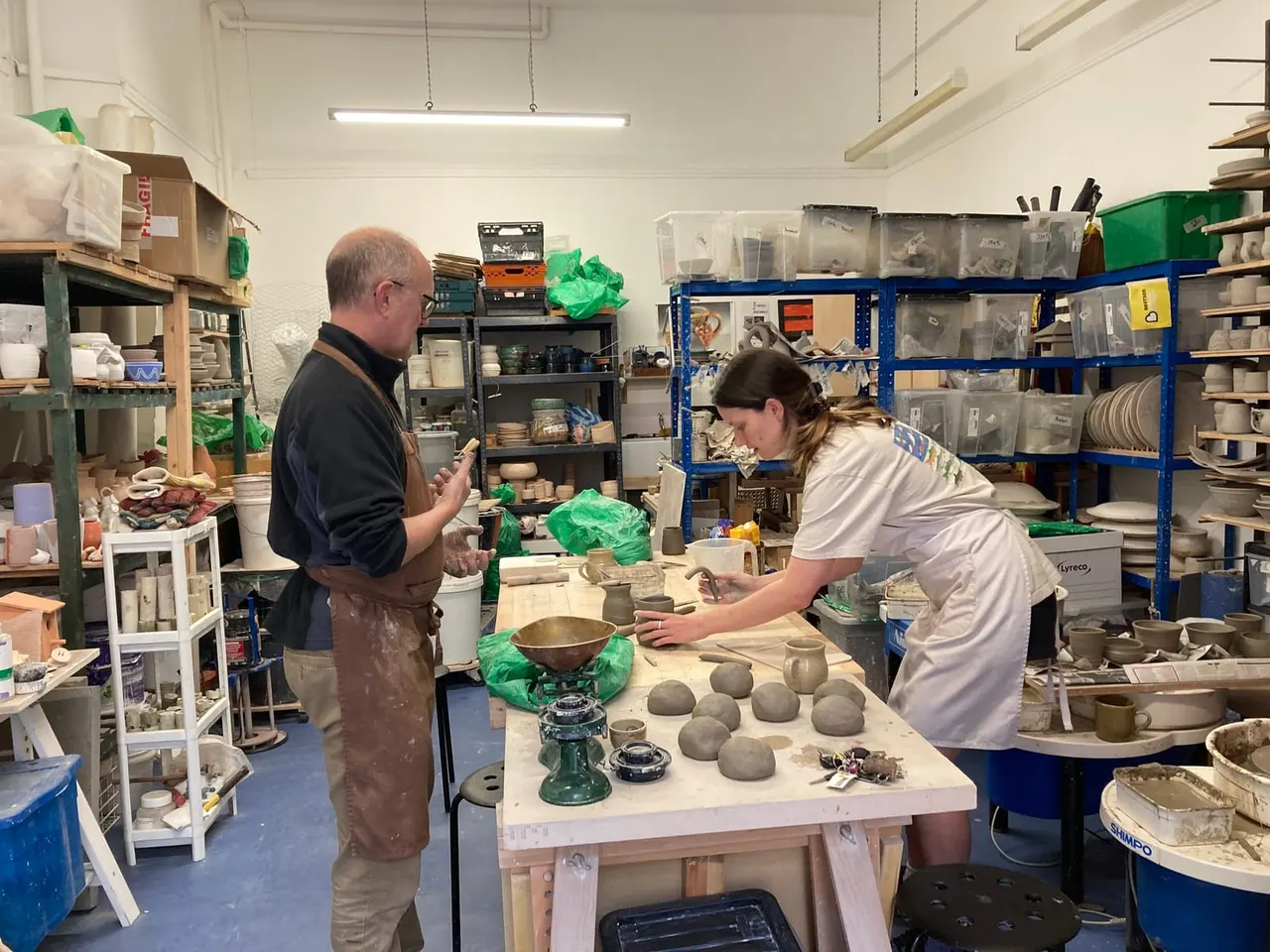 Two people working in a pottery studio surrounded by shelves of tools and materials, with clay pieces on a central work table.