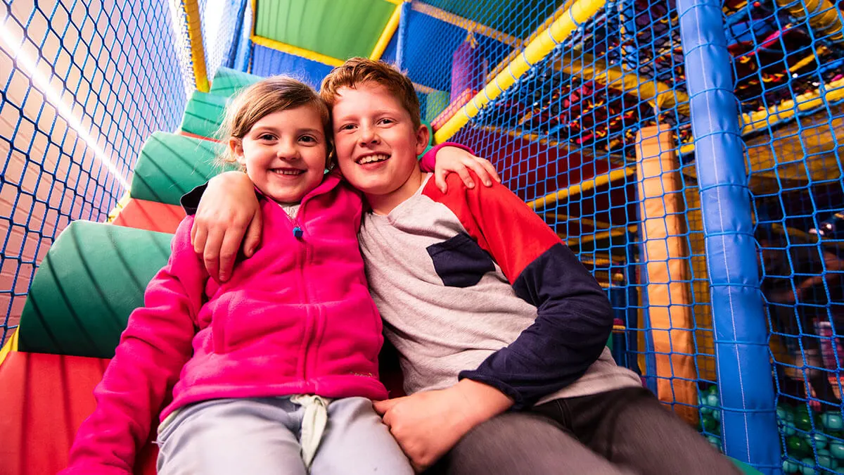 Two children smiling inside a colorful soft play structure with netting and padded equipment.