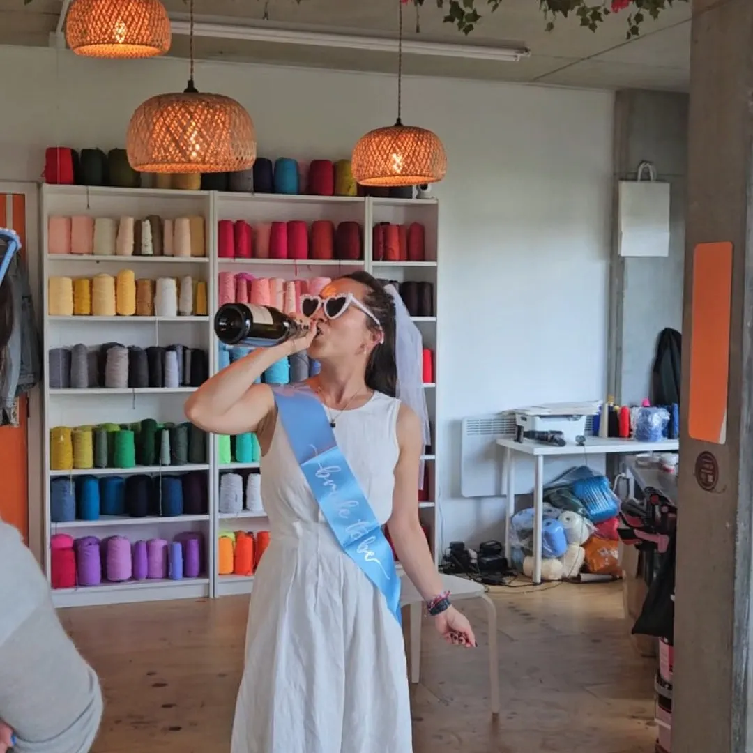 Woman in white dress and sunglasses drinking from a bottle in a craft studio with colourful thread shelving.
