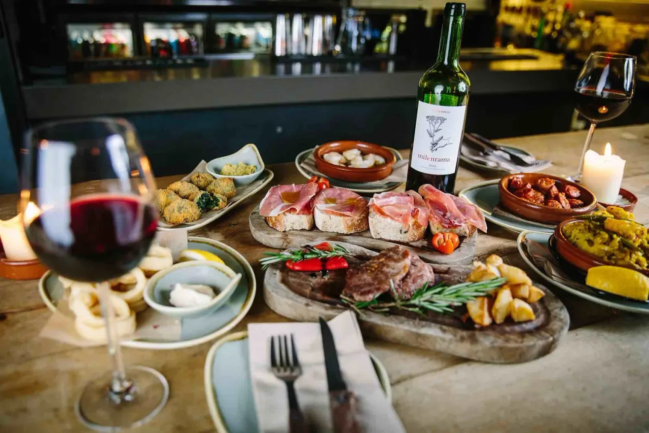 Table setting at a restaurant bar with Spanish tapas, cured meats, bread, wine bottles and glasses, and various small dishes.