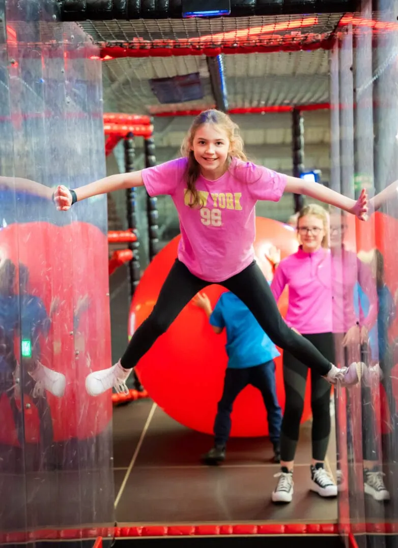Child in pink shirt jumping on trampoline with arms spread wide in indoor play facility with red and blue equipment.