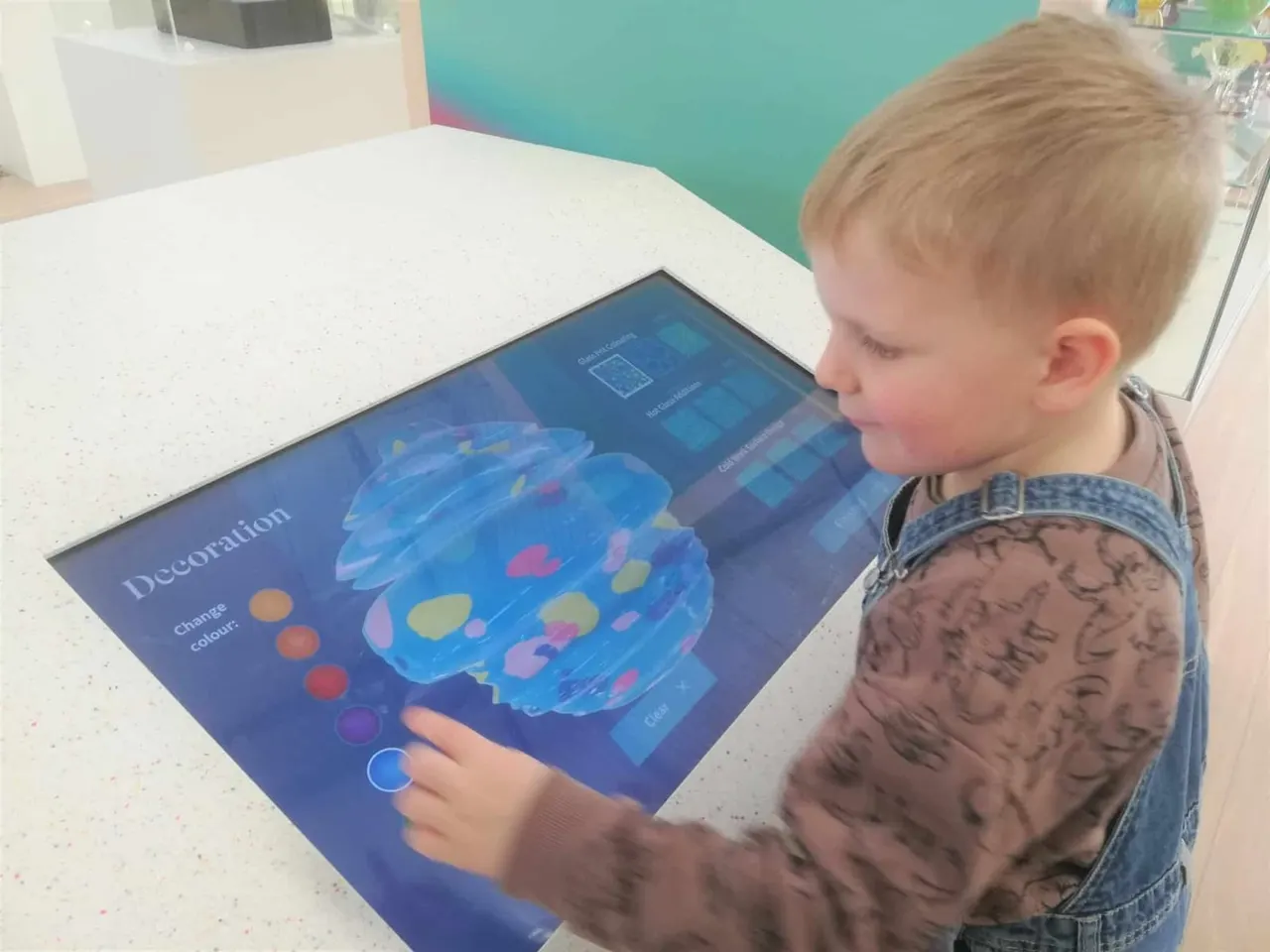 A young child interacts with a touchscreen display showing a blue decorative glass object with colour-changing options.