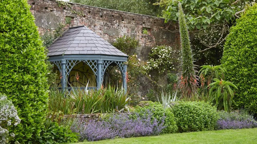Blue-painted gazebo with slate roof nestled in formal garden with hedges, lavender and flowering plants against stone wall.
