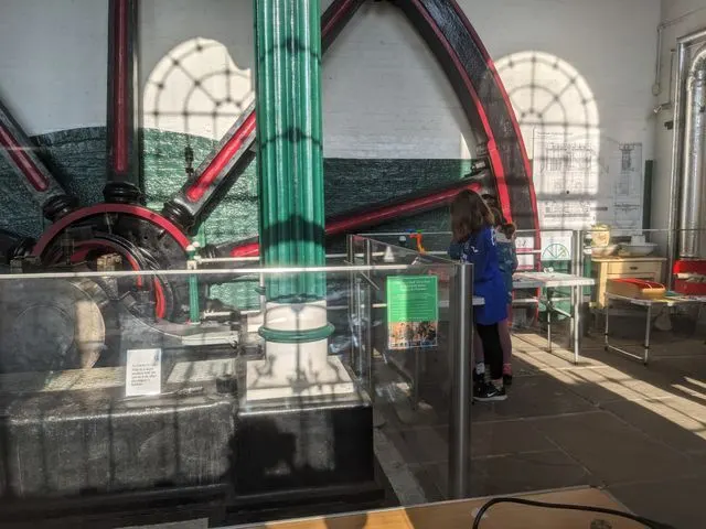 Visitor in museum gallery viewing large red and green industrial beam engine machinery behind glass display case.