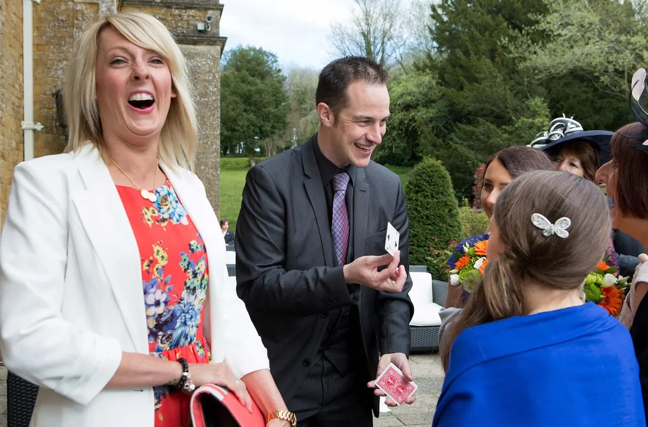 A magician in smart attire entertains guests at an outdoor event by a stone building, with children and adults gathered around watching.