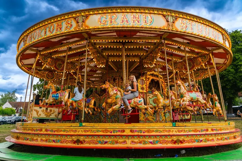 Ornate traditional carousel with illuminated horses and riders, set against a blue sky at a fairground.