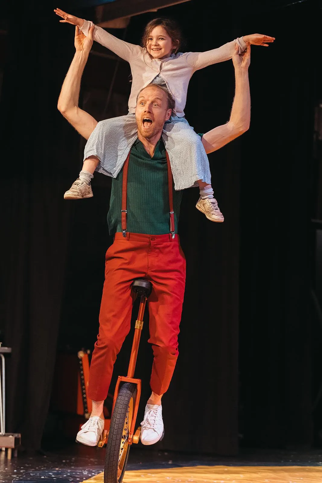 Adult performer riding a unicycle with a child balanced on his shoulders, both with arms outstretched in a circus act.