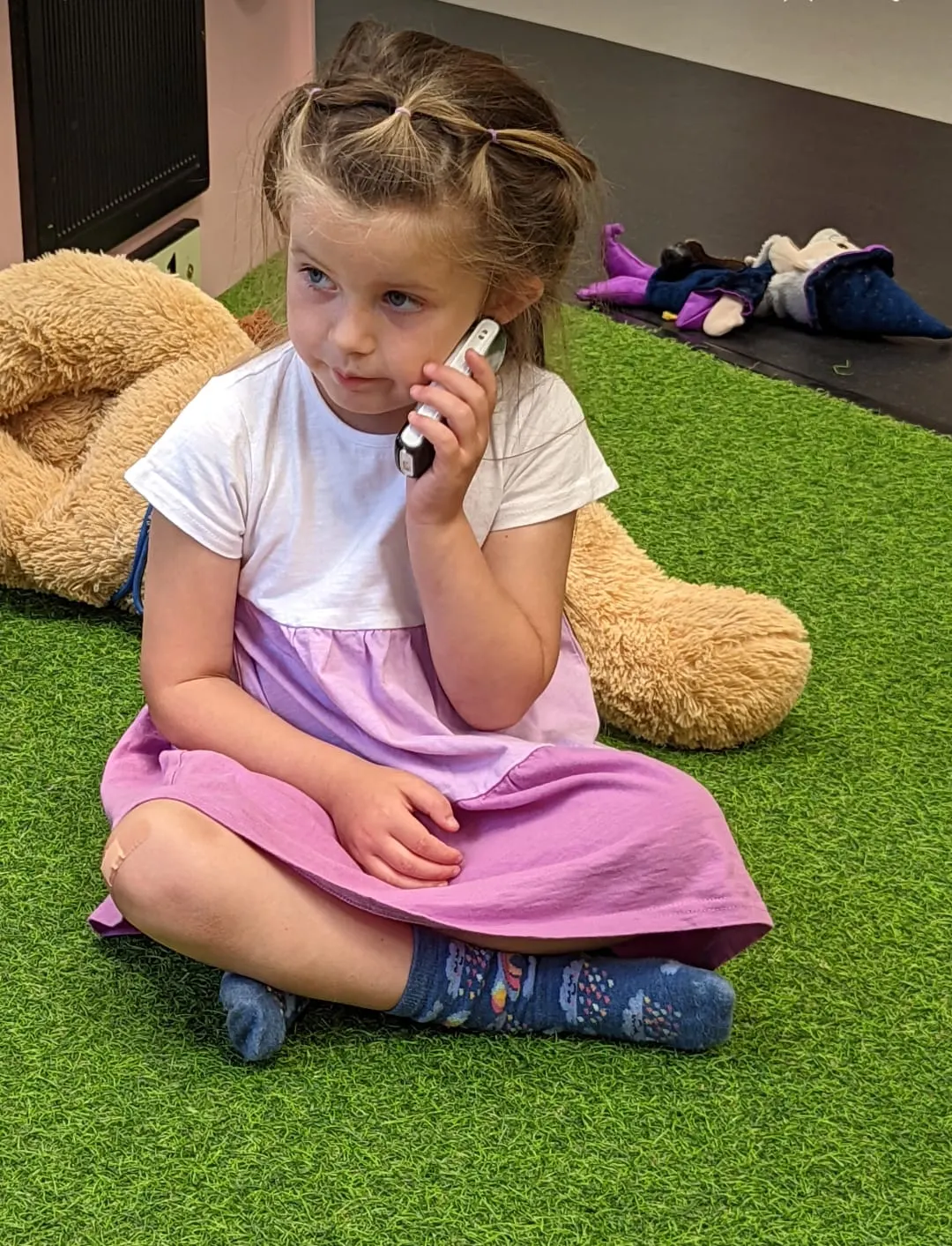 Young girl sitting on artificial grass next to a large teddy bear, holding a toy phone.