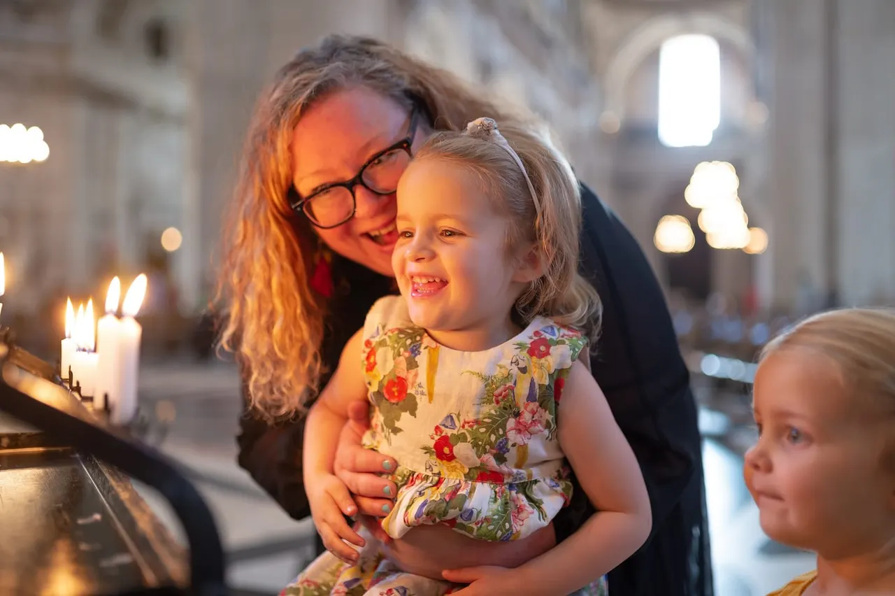 Adult and young child visiting a cathedral interior lit by candlelight, with stone architecture and glowing candles visible in the background.