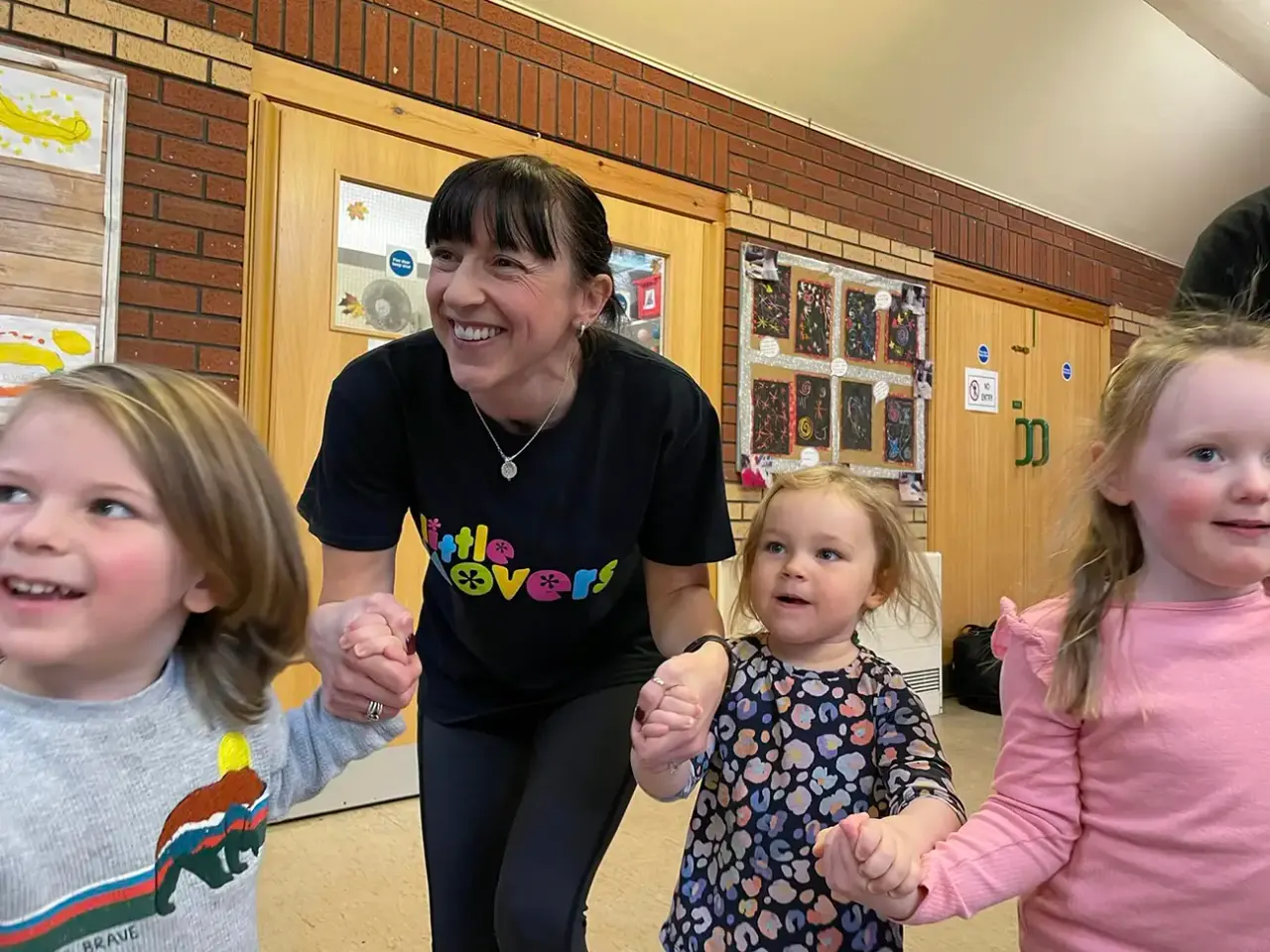 An adult staff member holds hands with three young children in an indoor activity space with brick walls and wooden doors.