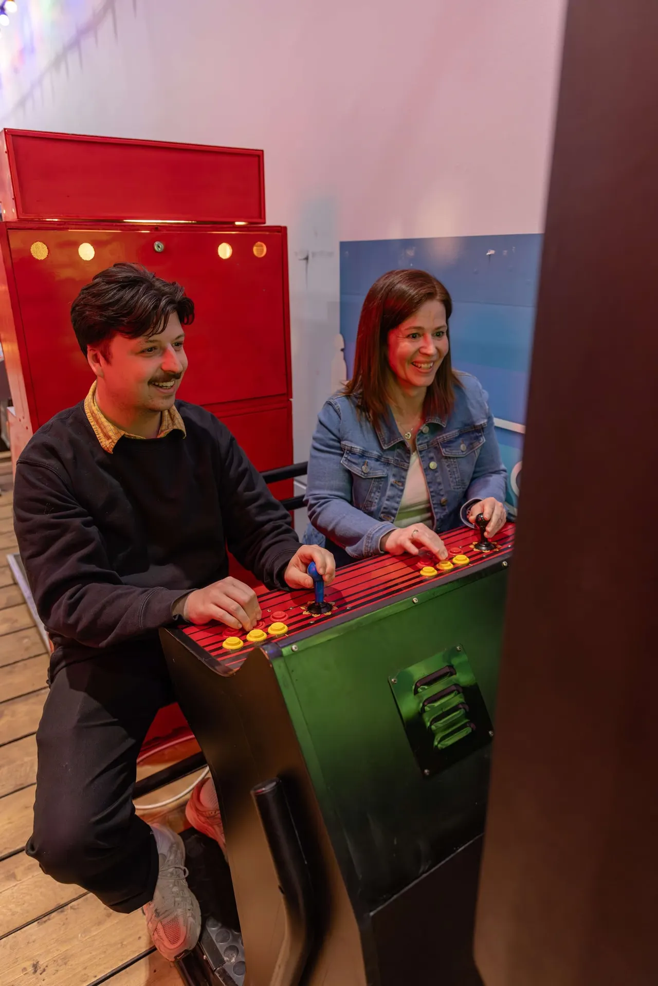 Two visitors playing a red and green tabletop game with illuminated buttons and controls at an interactive museum exhibit.
