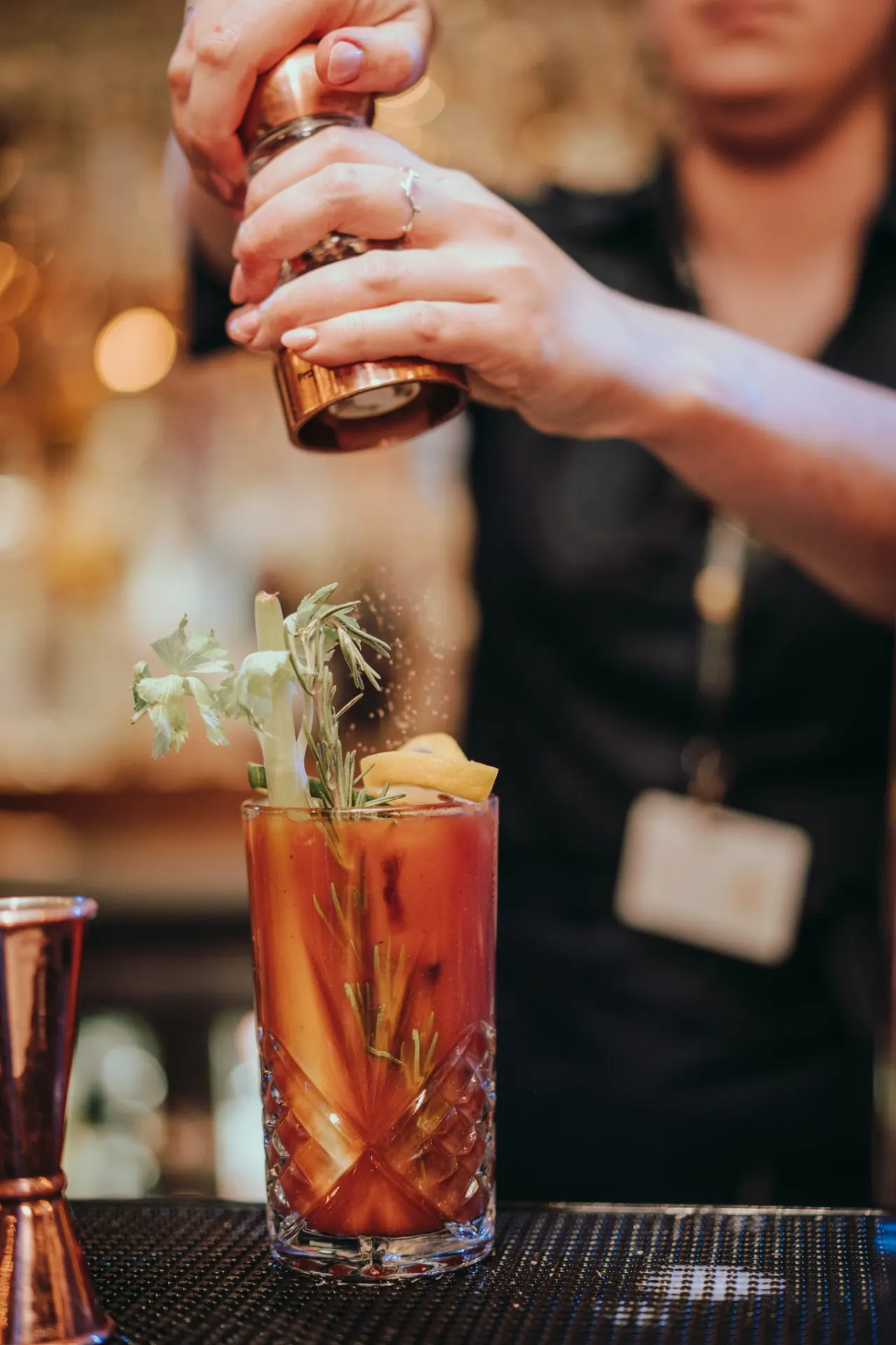Bartender pouring spirit into a craft cocktail garnished with rosemary and citrus at a bar counter.