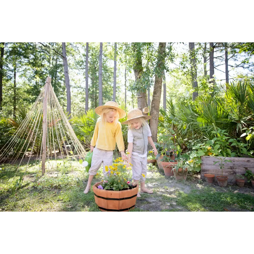 Two young children wearing straw hats explore a woodland garden with potted flowers and a rope teepee.