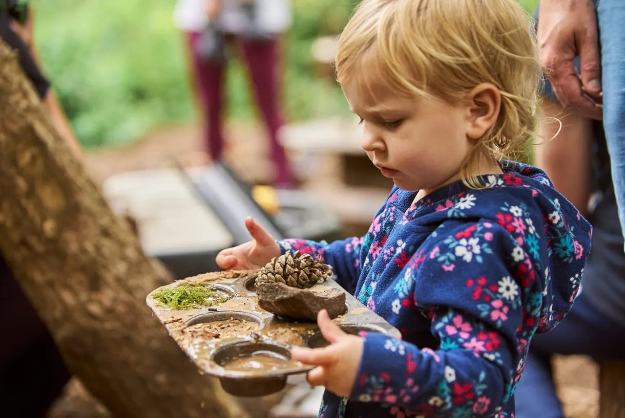 Young child in floral jacket touching natural materials and moss in outdoor woodland activity with adults nearby.