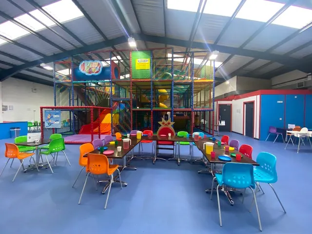 Colourful indoor play area with a large climbing structure, foam blocks, and bright chairs on a blue floor.