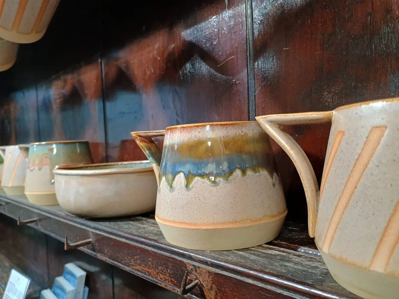 Handmade ceramic jugs and vessels with glazed finishes displayed on a wooden shelf behind glass.
