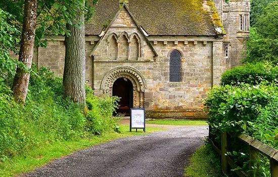 Stone chapel surrounded by trees and green grounds, with a curved arched doorway and information board at the entrance.
