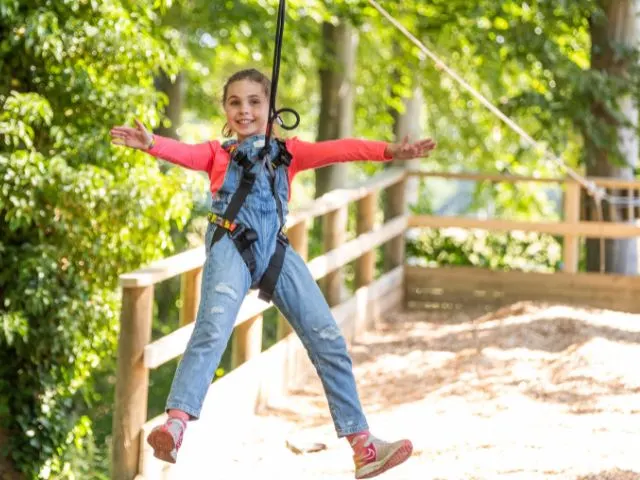 Child on a zip line with arms outstretched, suspended above a wooded adventure course.