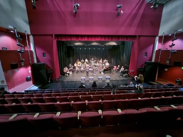 Orchestra performing on an illuminated stage with magenta walls, viewed from empty auditorium seating.