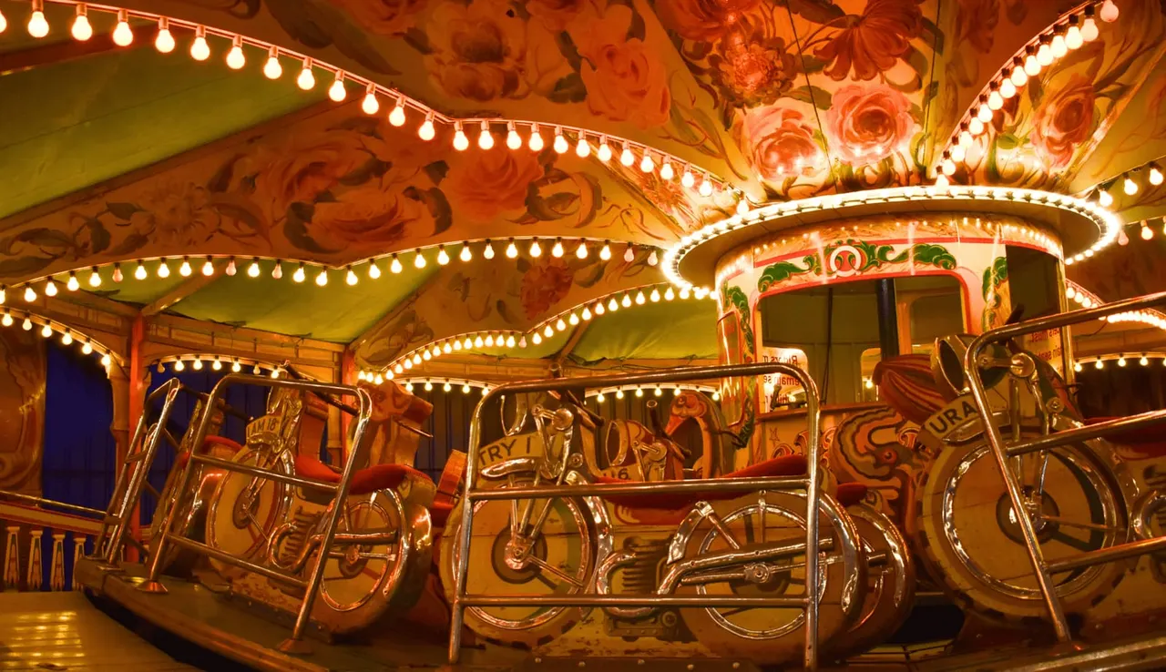 Brightly lit vintage carousel with ornate painted panels and decorative bulbs, featuring traditional fairground horses and vehicles.