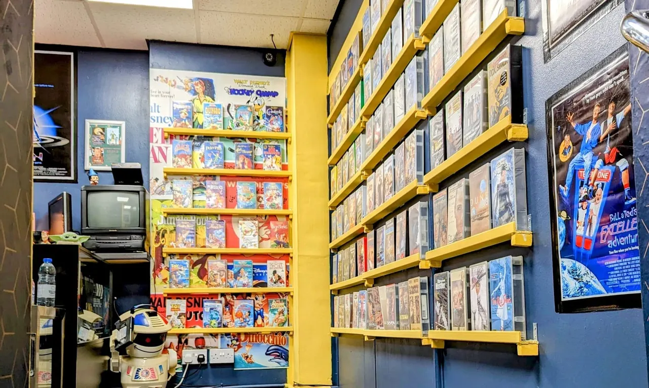 Interior of retro gaming arcade with yellow shelving displaying vintage video game boxes and memorabilia on blue walls.