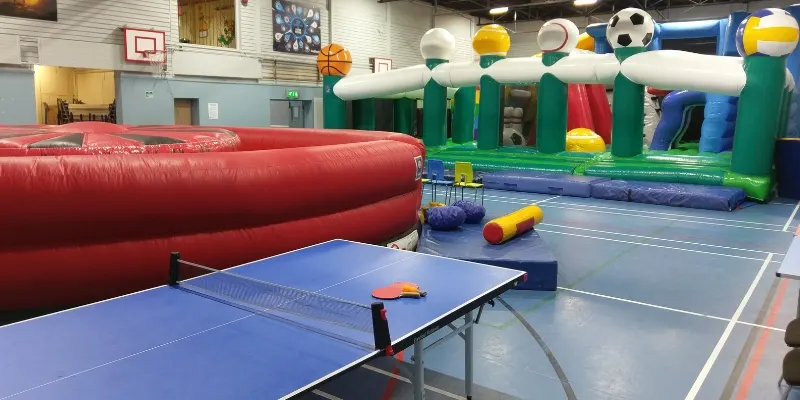 Indoor play area with table tennis table, large red inflatable, and colourful sports-themed bouncy castle.