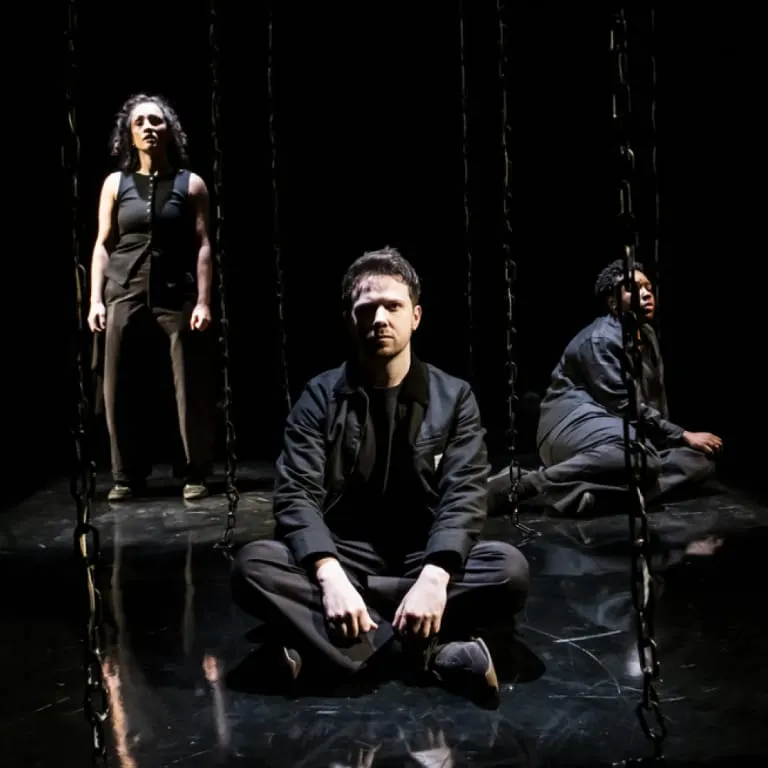 Three performers on a dark stage with hanging chains, two seated and one standing.