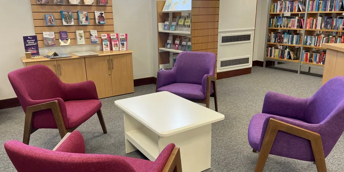 Library lounge area with colourful upholstered armchairs arranged around a white table, bookshelves and wooden storage units in background.