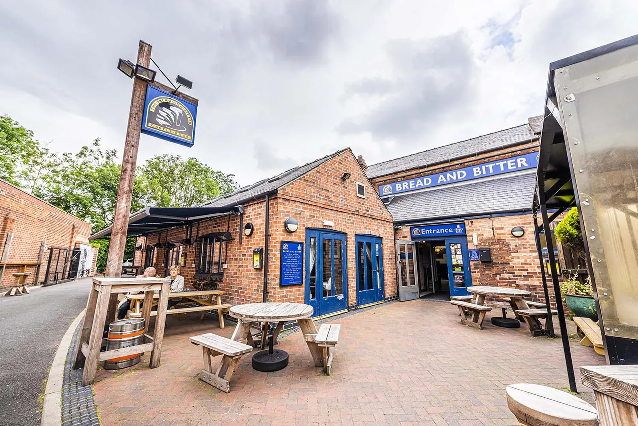 Exterior of a brick pub with blue doors and signage, outdoor seating with picnic tables, and a hanging pub sign, with a person seated outside.
